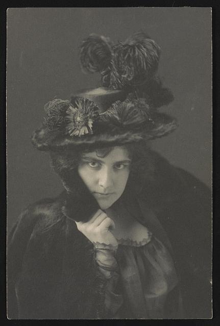 Black and white photo. A woman (Ethel Reed) looks towards the camera, draped in a fur coat and wearing a large, decorative hat with a large plume.