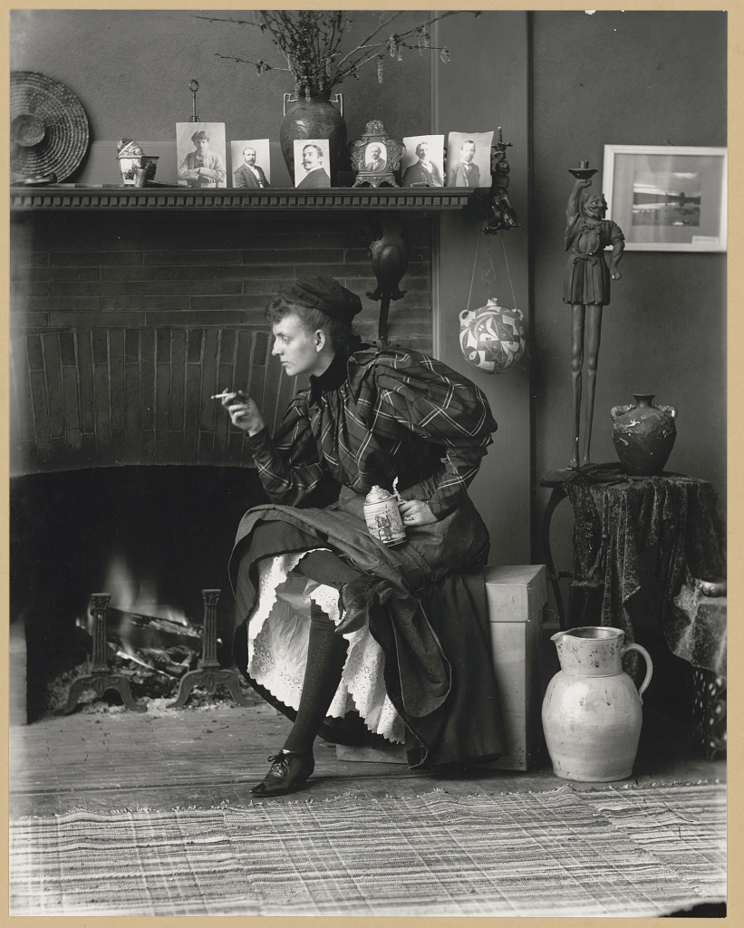 Black and white self portrait of Frances Benjamin Johnston. Frances is swearing a tartan dress and black stockings and a dark coloured cap. She is leaning left while holding a beer stein and a cigarette.
