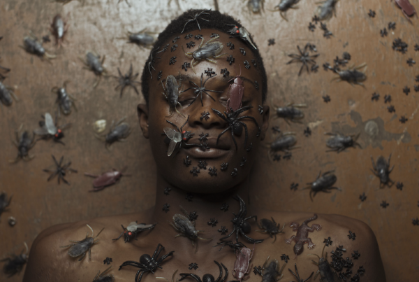Colour photograph. A person lies naked in their back, their face and the body (as well as the background) covered in plastic flies and insects. We see their head and shoulders only; their eyes are closed.