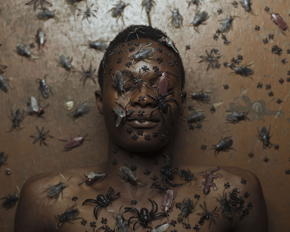 Colour photograph. A person lies naked in their back, their face and the body (as well as the background) covered in plastic flies and insects. We see their head and shoulders only; their eyes are closed.