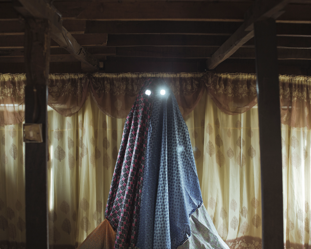 Colour photograph. A central figure is covered by a red and blue sheet, two white lights shining resembling eyes.