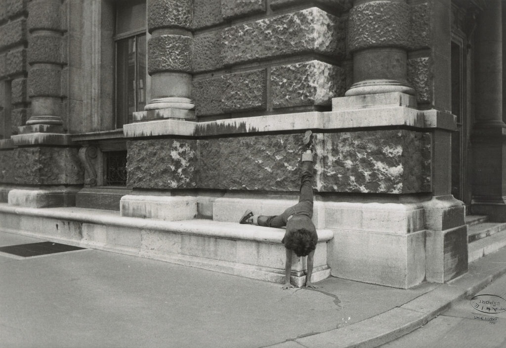 Black and white photo. A person stands on their hands with their legs against a brick wall - one horizontal, one vertical.