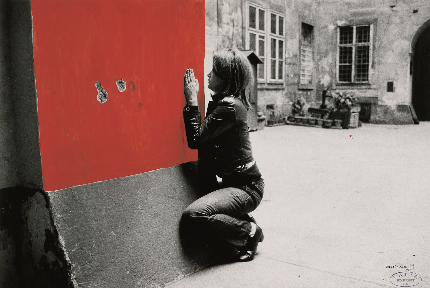 Black and white photo. A woman kneels next to a wall. The photograph of the wall section has been painted red.