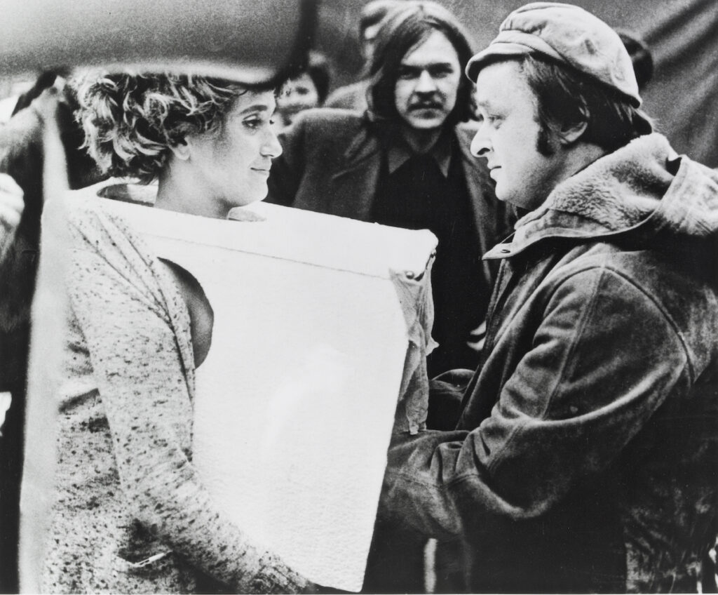 Black and white photo depicting a woman (VALIE) wearing a box around her torso. Opposite her, a man reaches his hands into the box while maintaining eye contact.