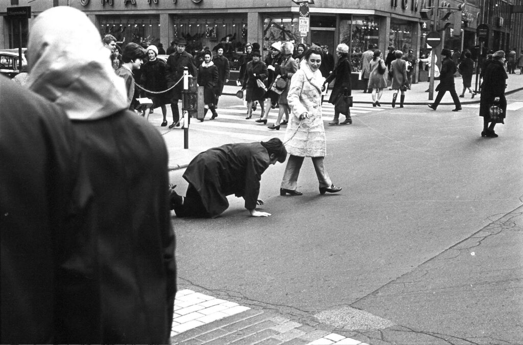 Black and white photograph of a woman leading a man on a lead though some city streets. He is crawling after her.