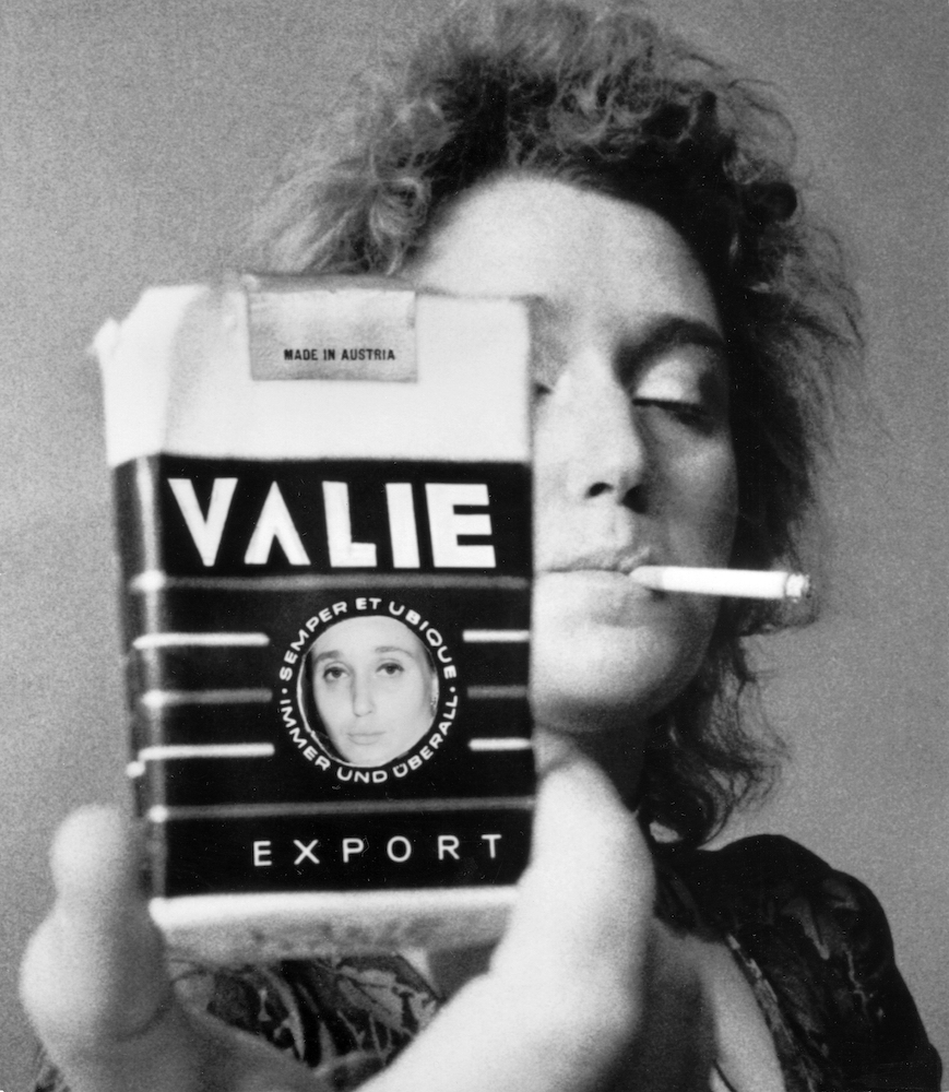 Black and white portrait of VALIE holding a cigarette packet bearing her name. She holds a cigarette in her mouth.