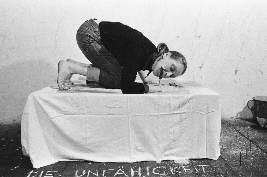 Black and white photo. A woman kneels on a table, which is covered with a white cloth. In her mouth, she is holding some kind of tool, with which she is working.