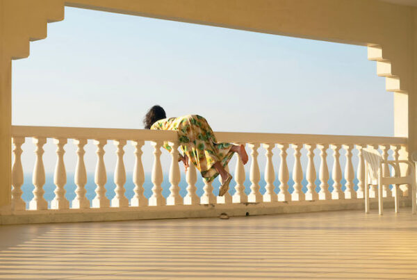 Colour photograph. A woman leans over a white railing; we see her from behind, several metres away. One of her feet is bare; on the other there is a sandal shaped like a fish.