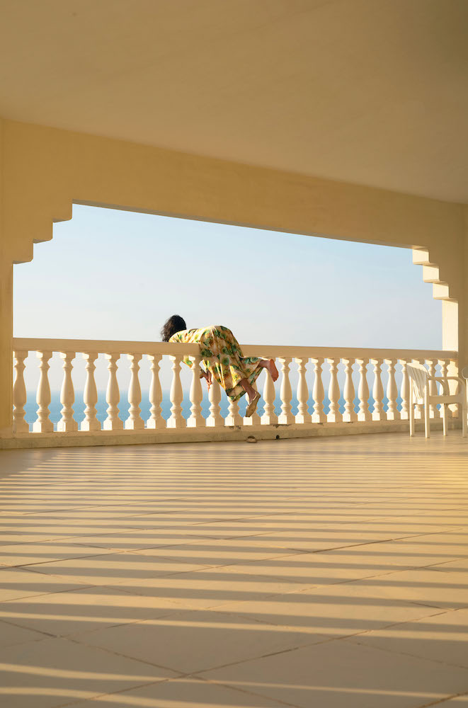 Colour photograph. A woman leans over a white railing; we see her from behind, several metres away. One of her feet is bare; on the other there is a sandal shaped like a fish.