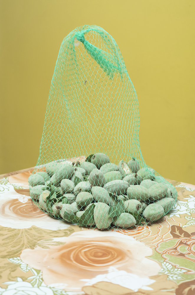 Colour photograph. A green mesh bag of green almonds sits on a tablecloth patterned with orange flowers, in front of a yellow wall.