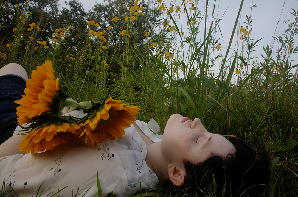 Alex lies in a patch of long grass, holding a bouquet of sunflowers.
