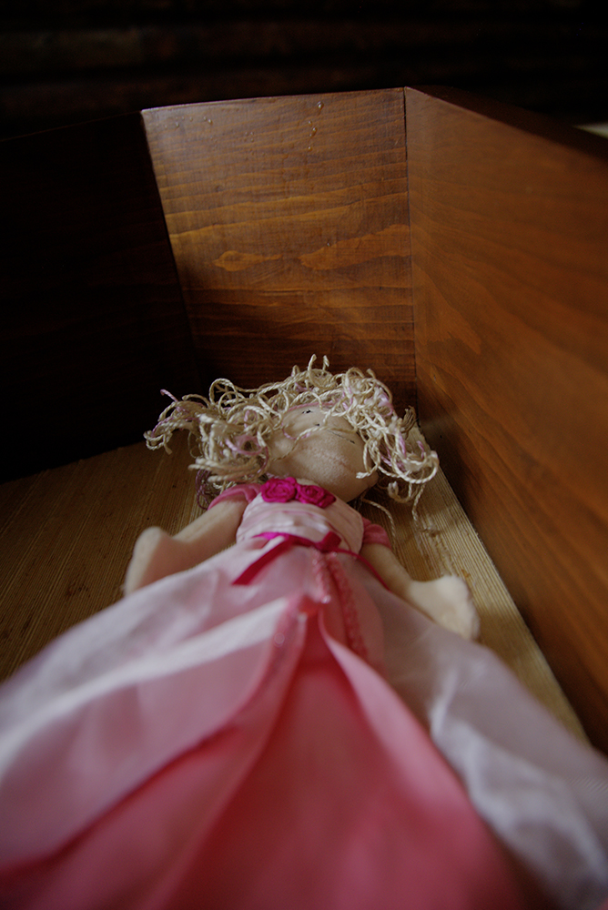 A soft toy doll wearing a pink dress, lying in a wooden drawer.