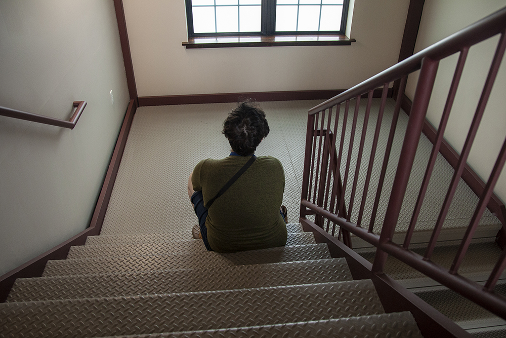 Alex faces away from us, sitting on metal steps with a metal handrail. An interior, possibly a stairwell.