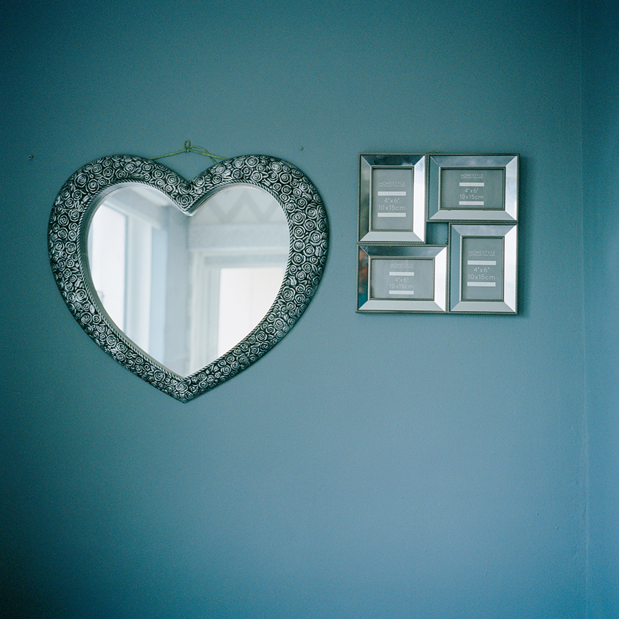 Colour photograph of a heart shaped mirror and empty photograph frames on a blue wall.