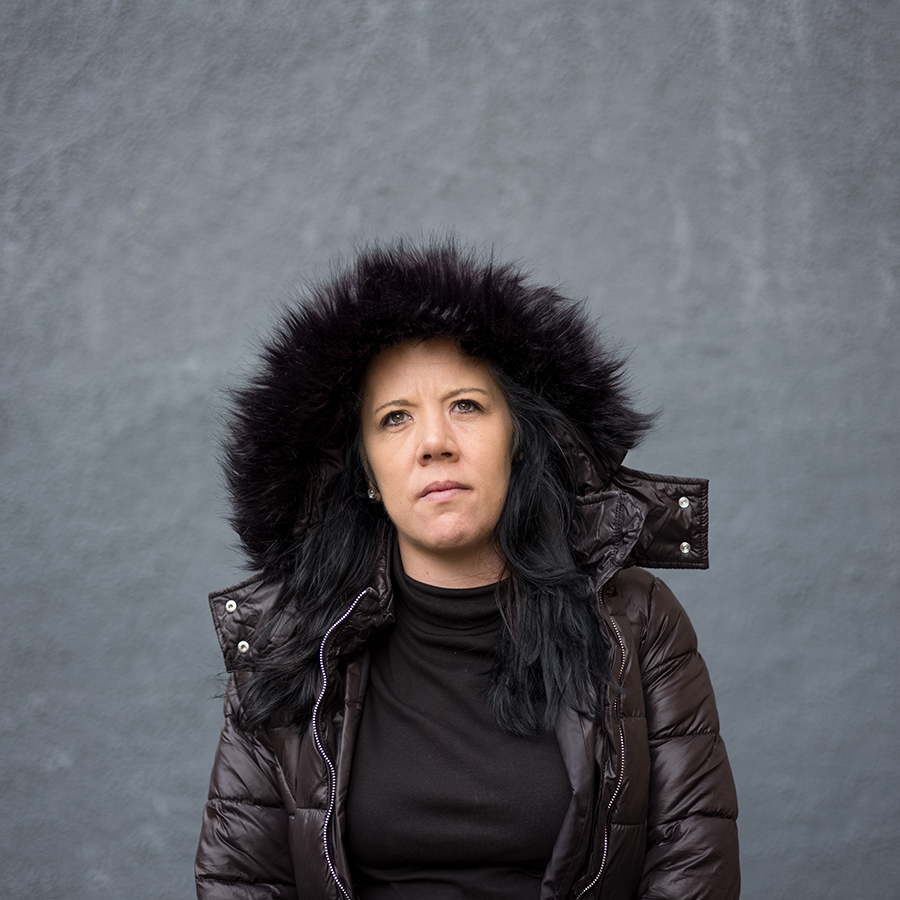 Colour photograph. Lyndsey looks beyond the camera, wearing a fur lined parka. She stands against a grey wall.