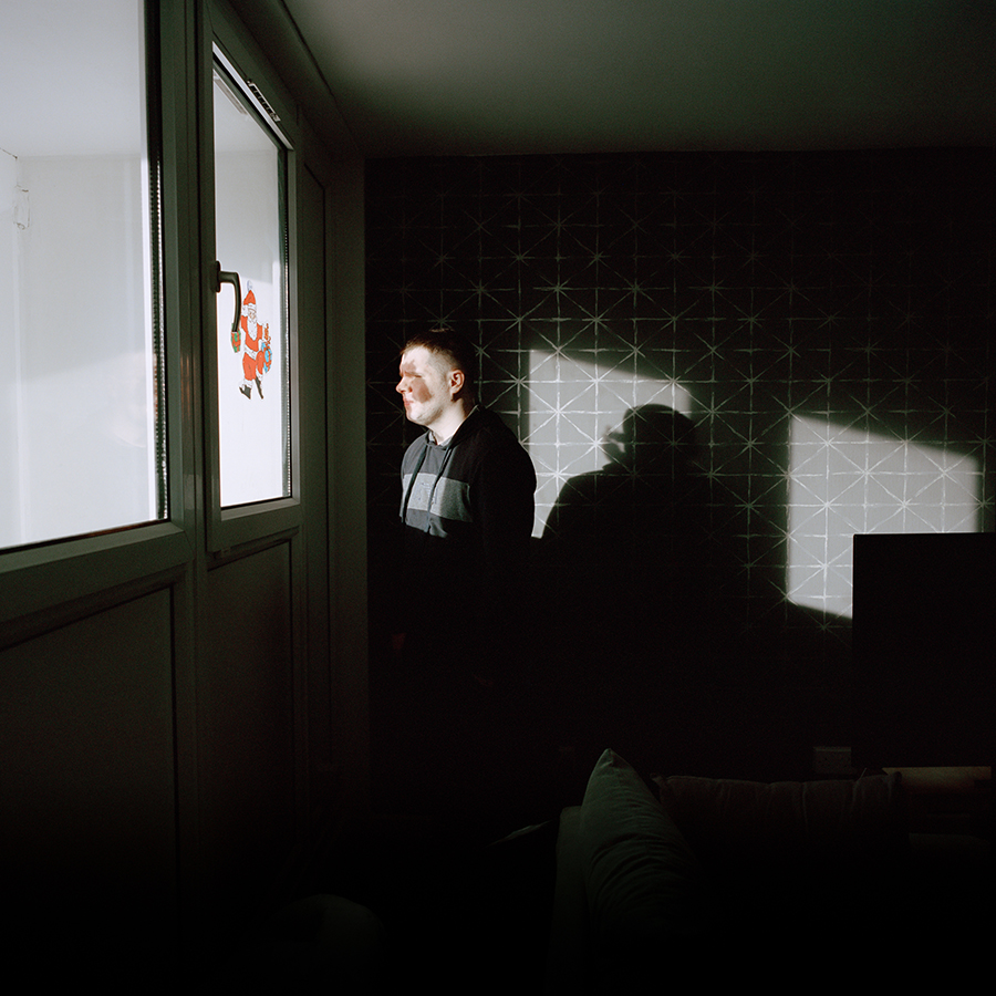 Colour photograph. A heavily darkened room. Marcus looks left, though a window, the only point of light. There is a Santa Claus sticker on the window.