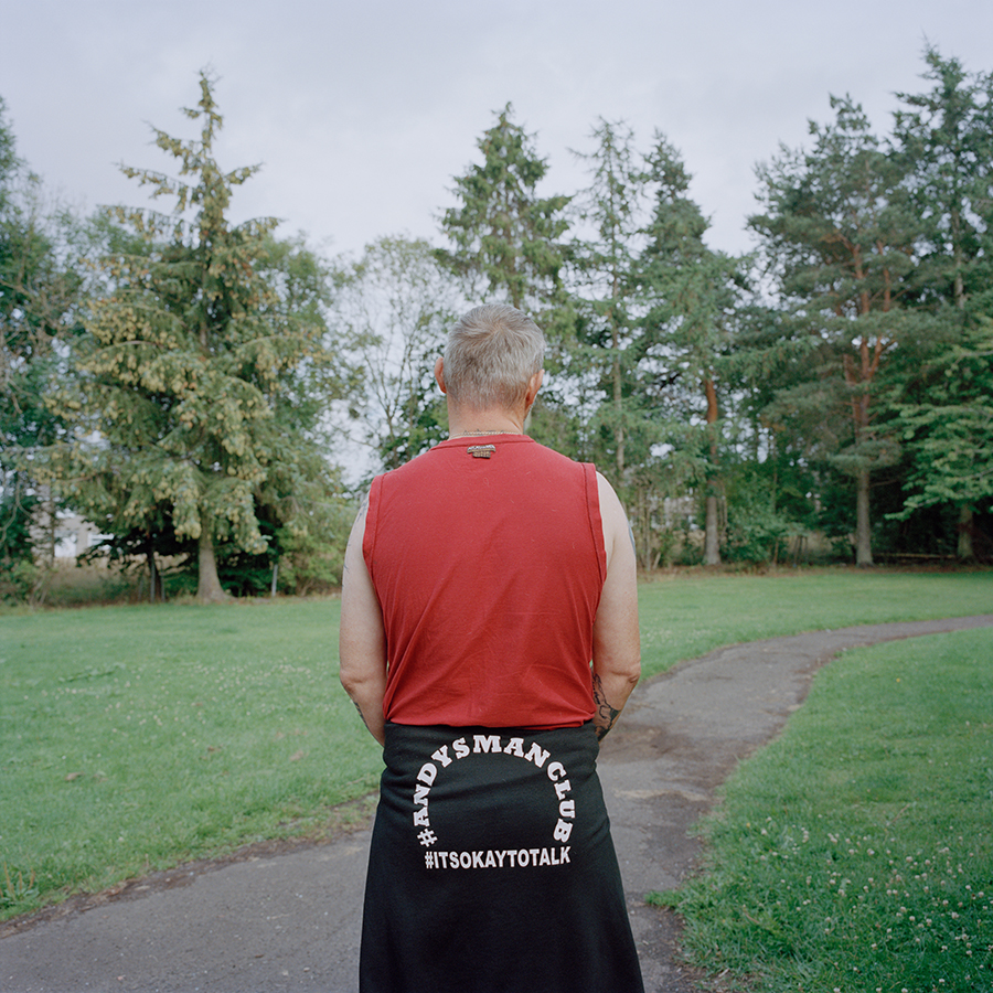 Colour photograph. Mark stands in a park, facing away from us into the treeline. He wears a red tank top and has a black top tied around his waist which reads '#AndysManClub #ItsOkayToTalk'.