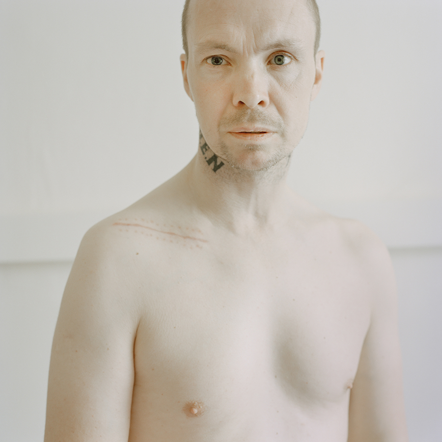 Colour photograph. Michael faces the camera. He is not wearing a shirt. The E.N. of his 'E.D.E.N.' neck tattoo is visible, alongside a long, faded scar on his shoulder.