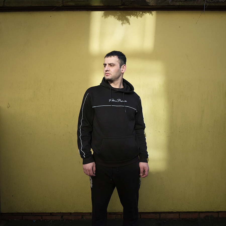 Colour photograph. A person stands in front of a yellow wall, wearing a black tracksuit.