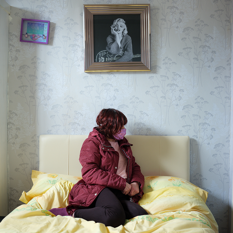 Colour photograph. A person in a red coat sits on a bed, wearing a pink face mask. Their face is turned to the right of the image. On the wall above them, we see a framed photograph of Marilyn Monroe.