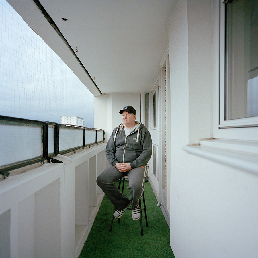 Colour photograph. Tam sits in a stool on a balcony comprised of narrow white walls and a green carpet. He wears a grey tracksuit and a black baseball cap. His hands are folded in his lap.