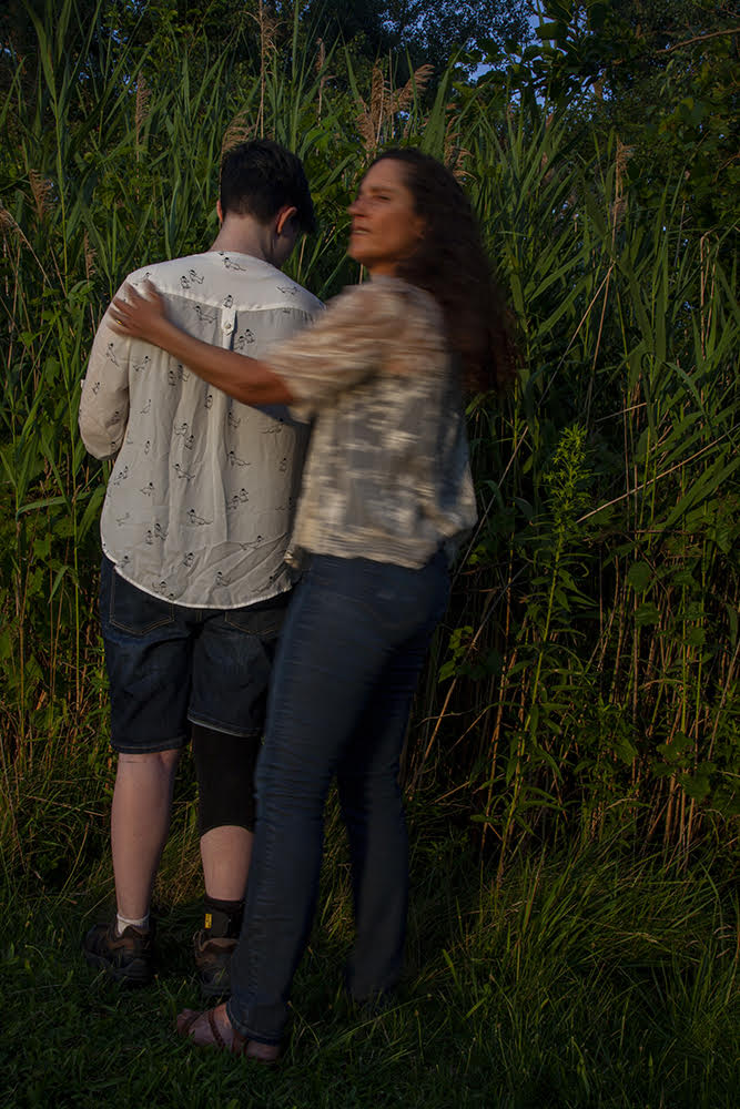 Alex stands among tall grass, facing away from us. A woman stands at their side, with her arm around Alex's shoulders.