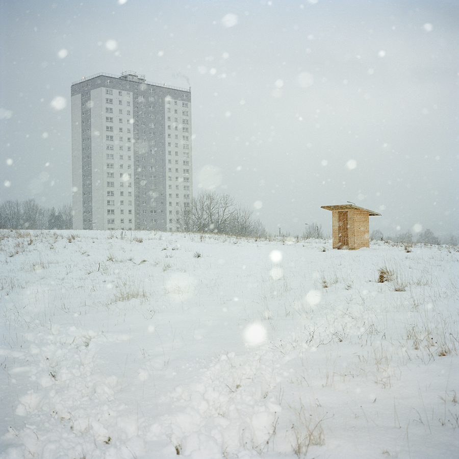 Colour photograph. A block of flats and a small wooden hut amid a snowy landscape.