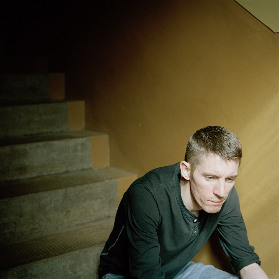 Colour photograph. Andrew sits on concrete stairs, in front of a wall painted yellow.