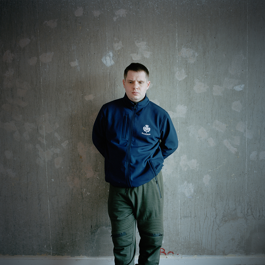 Colour photograph. Marcus stands against an undecorated wall stained with drips. He wears a navy blue jacket and green trousers.