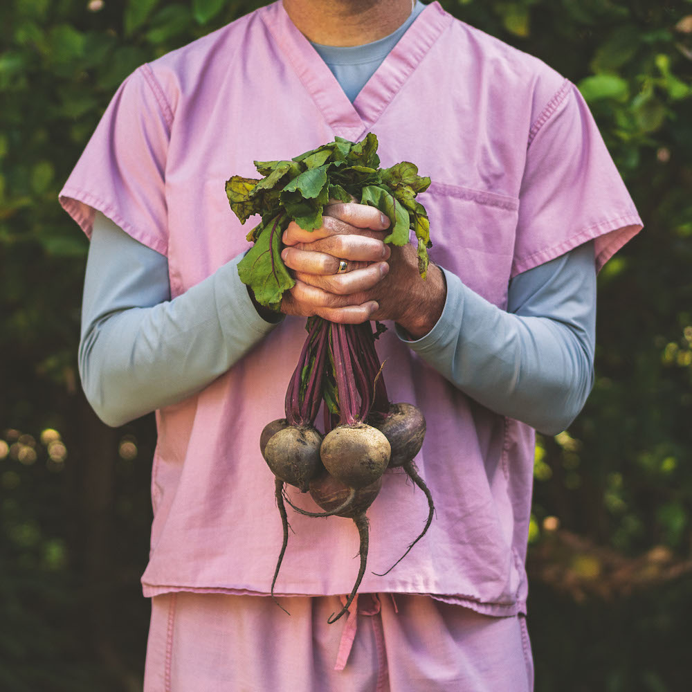 Colour photograph. A person wearing a pink (possibly medical) uniform holds a bundle of turnips; only their torso and hands are visible.