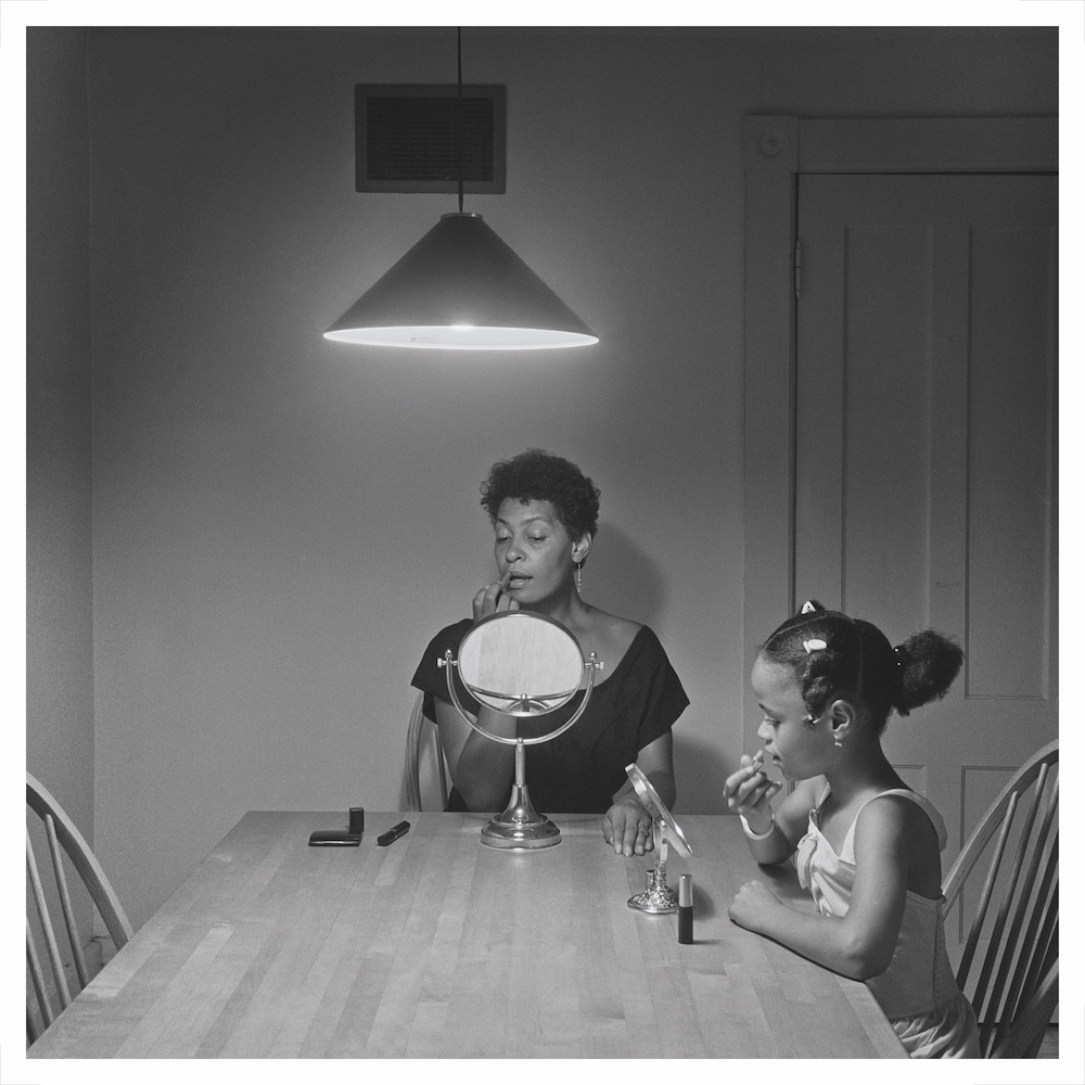 Black and white photograph. A woman and child sit at a kitchen table, applying lipstick.