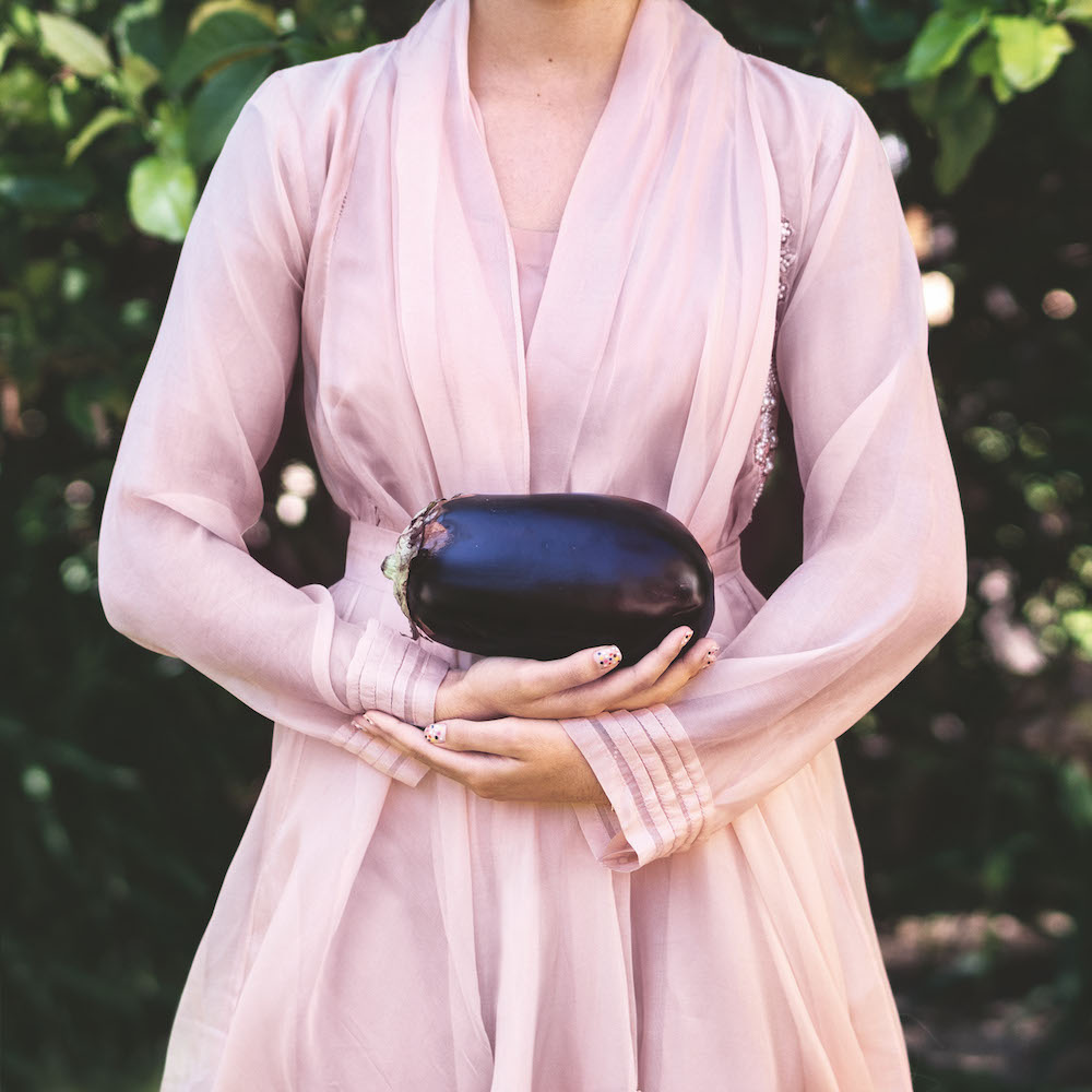 Colour photograph. A person in a light pink dress holds a large aubergine.