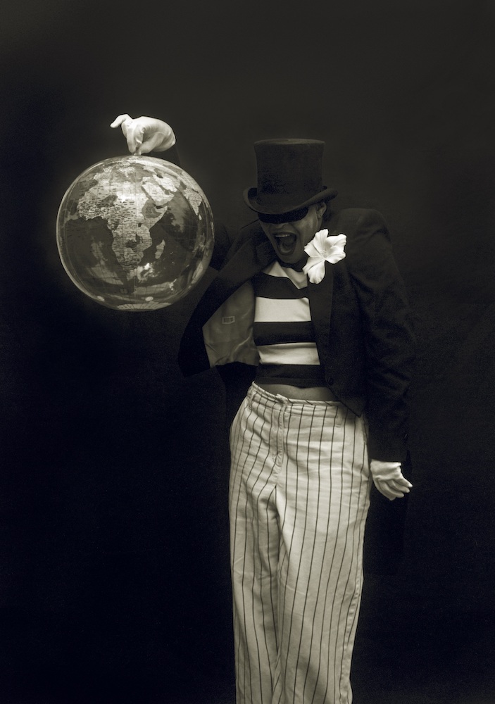 Black and white photograph. A person in a top hat, striped black and white top and black jacket, and white pinstripe trousers holds up a globe in a gloved hand.