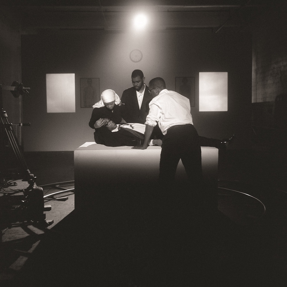 Black and white image. Two men and a woman lie another man down upon a central platform, looking on with concern.