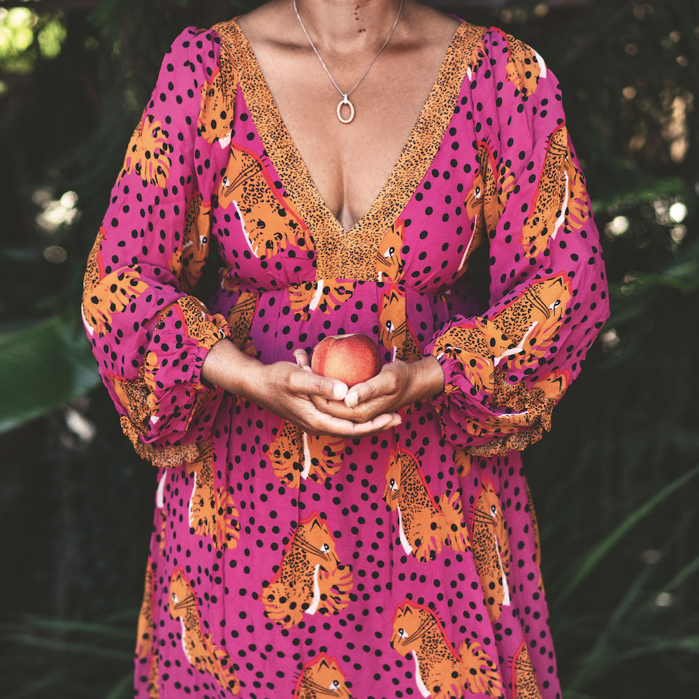 Colour photograph. A person in a pink dress, patterned with black spots and a leopard design, holds a ripe peach.