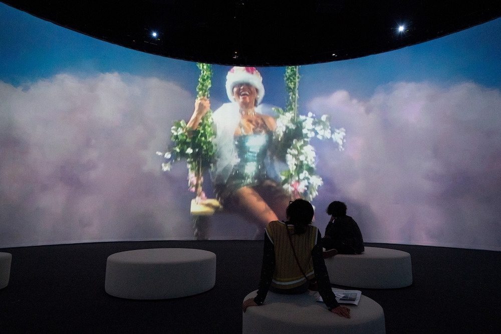 Colour photograph. People sit on fabric seats in a darkened room, in which a projection of Carrie's film is playing. We see a woman in a crown, swinging on a swing in front of a backdrop of clouds.