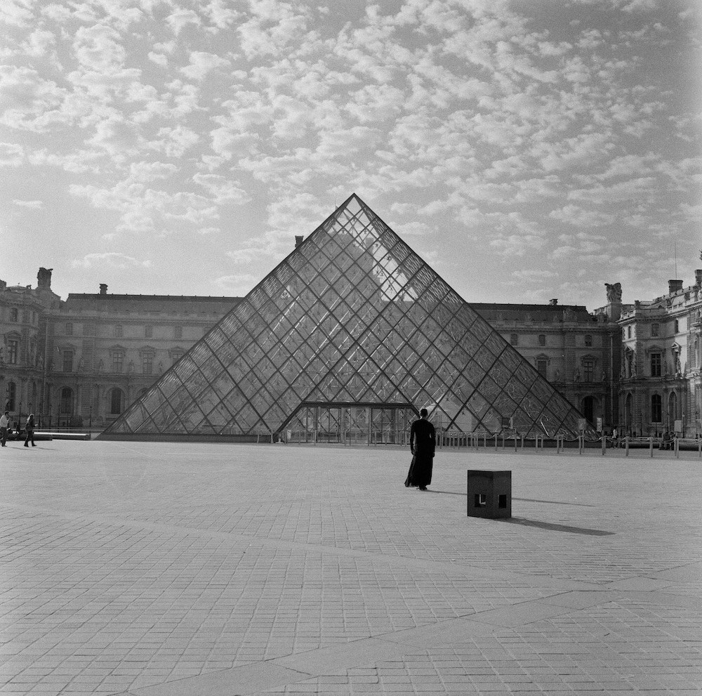 Black and white photograph. A person wearing a long, dark coloured dress stands in front of The Louvre, their back turned towards the camera.