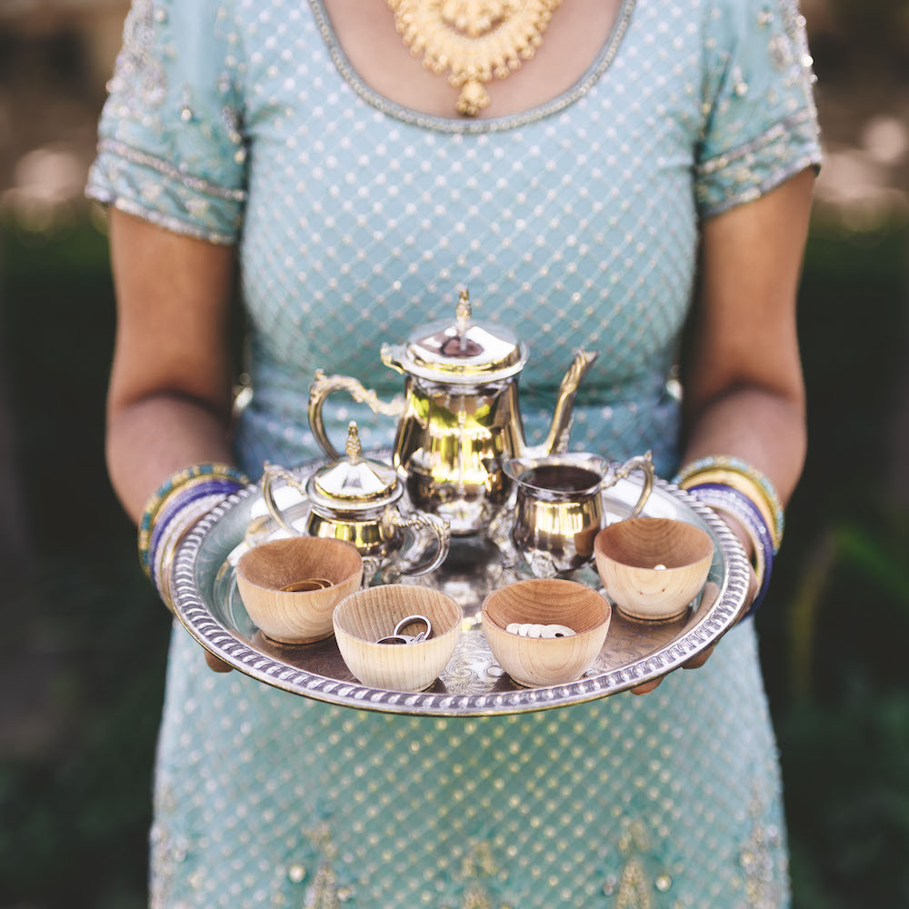 Colour photograph. A person wearing a light blue dress embroidered with a gold pattern holds a silver tray. On the tray is a silver tea set with small wooden cups.