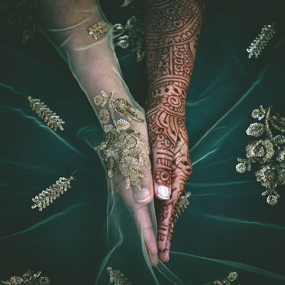 Colour photograph. Two hands are held together, one covered with a translucent green fabric embroidered with gold plants. The other hand is decorated with a detailed henna pattern.
