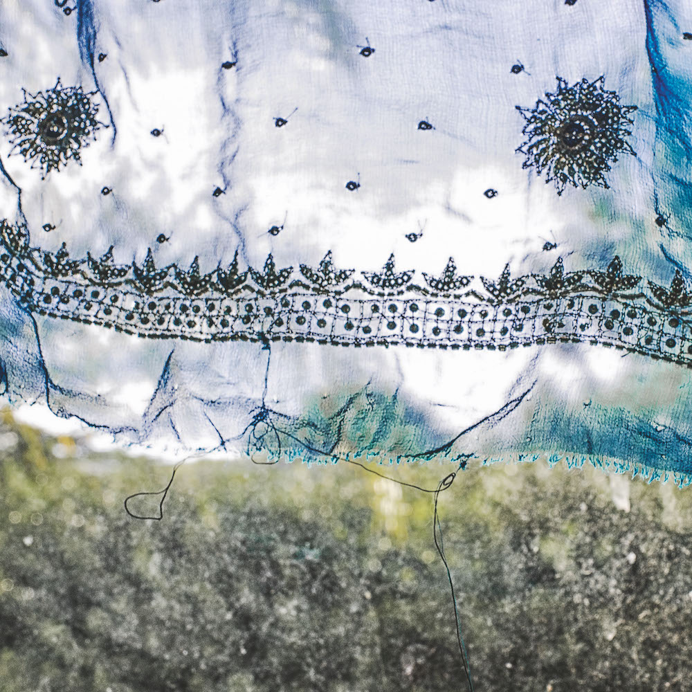 Colour photograph. White fabric with blue embroidery hangs in front of a green background.