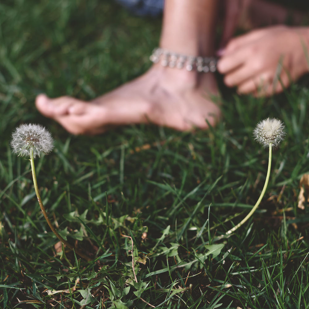 Colour photograph. A bare foot with an ankle bracelet steps onto grass. There are some dandelion seed heads in the foreground.