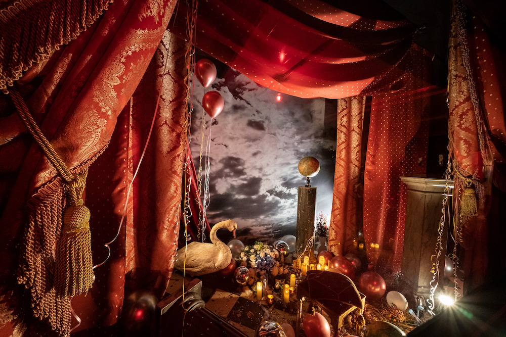 Colour photograph. Red curtains create a proscenium which surrounds an array of objects, such as a taxidermy swan, candles, and red balloons.