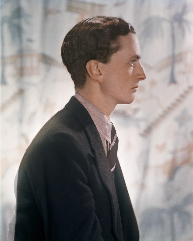 Colour photograph. Edward James faces to the left, wearing a black suit and pink shirt. The background is a piece of white fabric dedicated with pale blue palm trees.