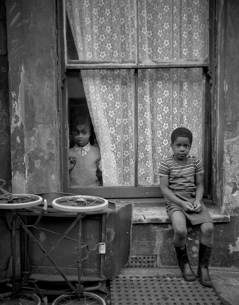 Black and white photograph. A child sits on a windowsill near an upturned pram. Another child looks out of the window behind them.