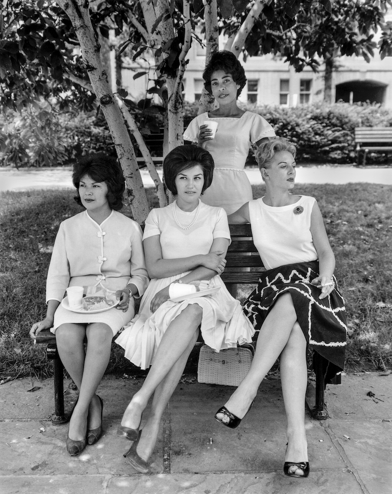 Black and white photograph. Three women sit on a bench in a park, while another woman stands behind them.