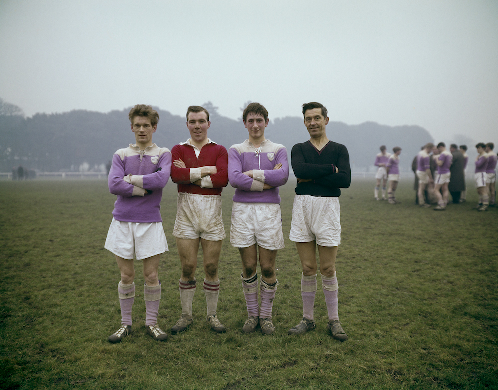 Colour photograph. Four men in sports kit (one in red, one in black, and two in purple) stand on a field. Other team members occupy the background.