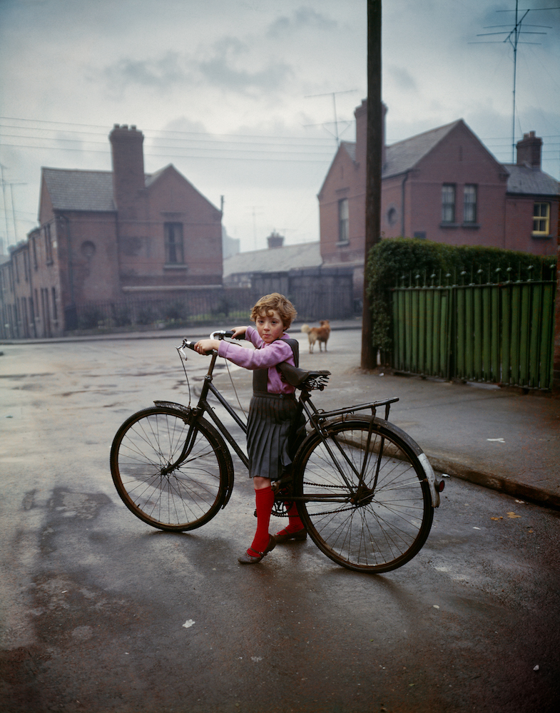 Colour photograph. A young girl in a pinafore dress stands in the street with a bicycle.