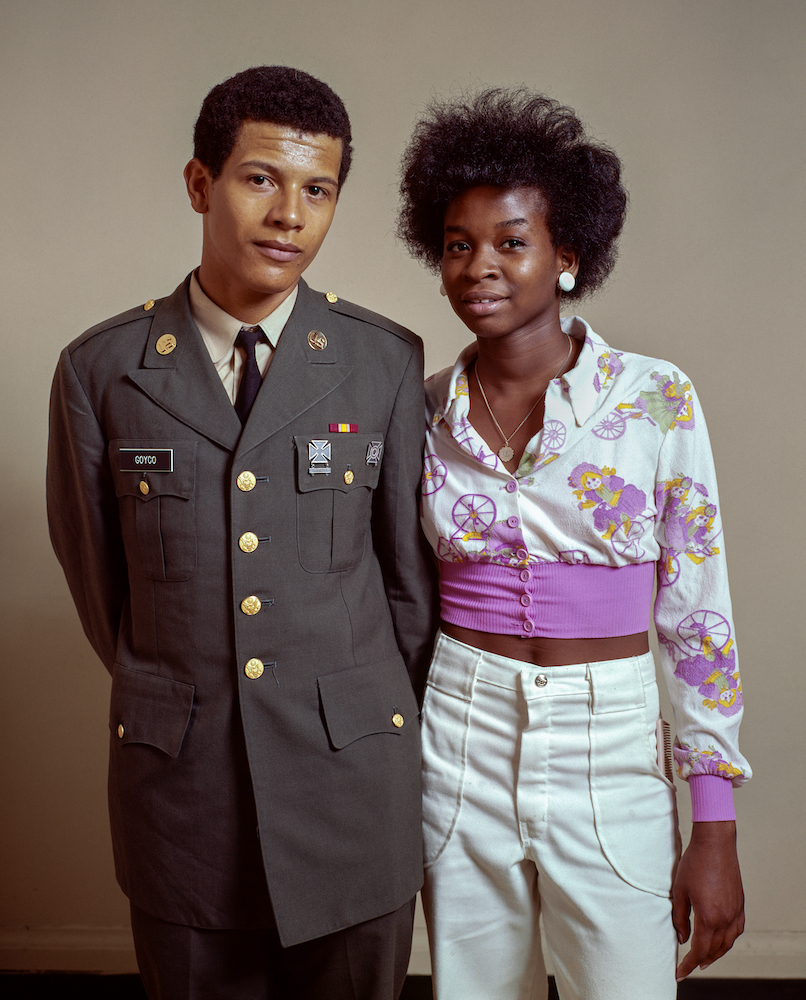 Colour photograph of a man and woman standing together. The man wears a military uniform, while the woman is dressed in a white and pink shirt and white trousers.