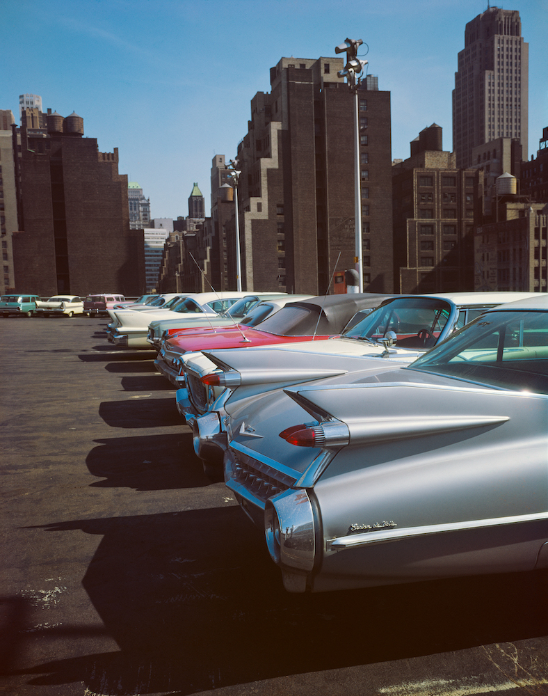 Colour photograph. A row of cars lined up in a car park. There are tall buildings in the background.