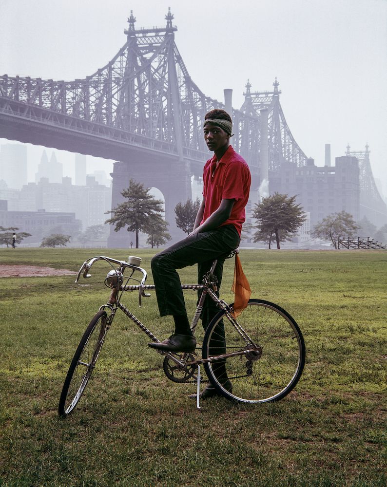 Colour photograph of a man sitting on a bicycle. Queensboro Bridge is visible in the background.